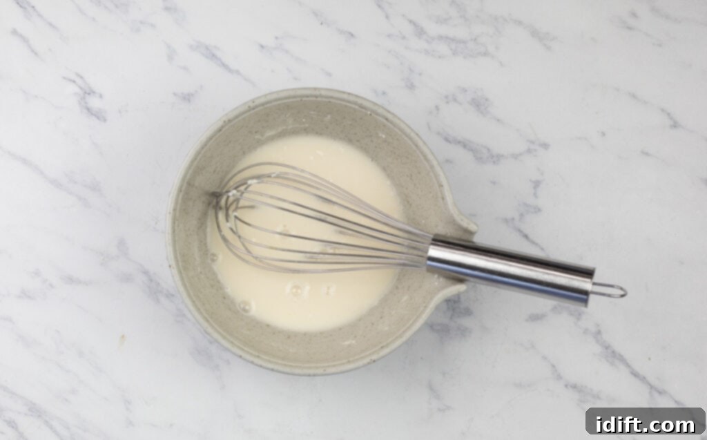 A bowl containing a whisk and a smooth, light-colored slurry, prepared for thickening the pan gravy, resting on a white marble surface.