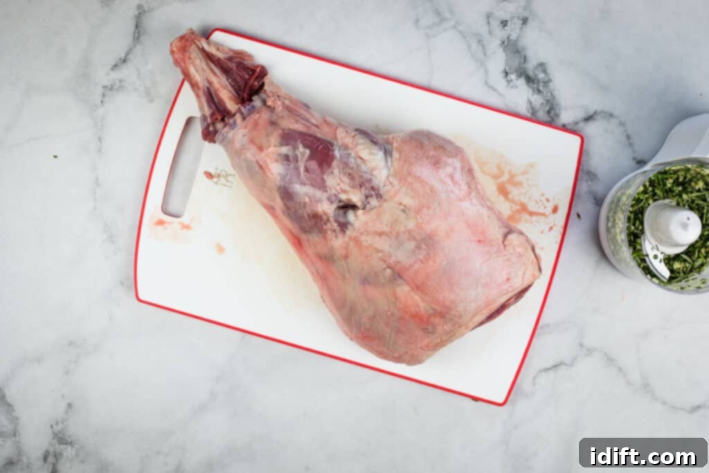 A leg of lamb on a cutting board, with a knife making an initial cut around the bone for frenching.