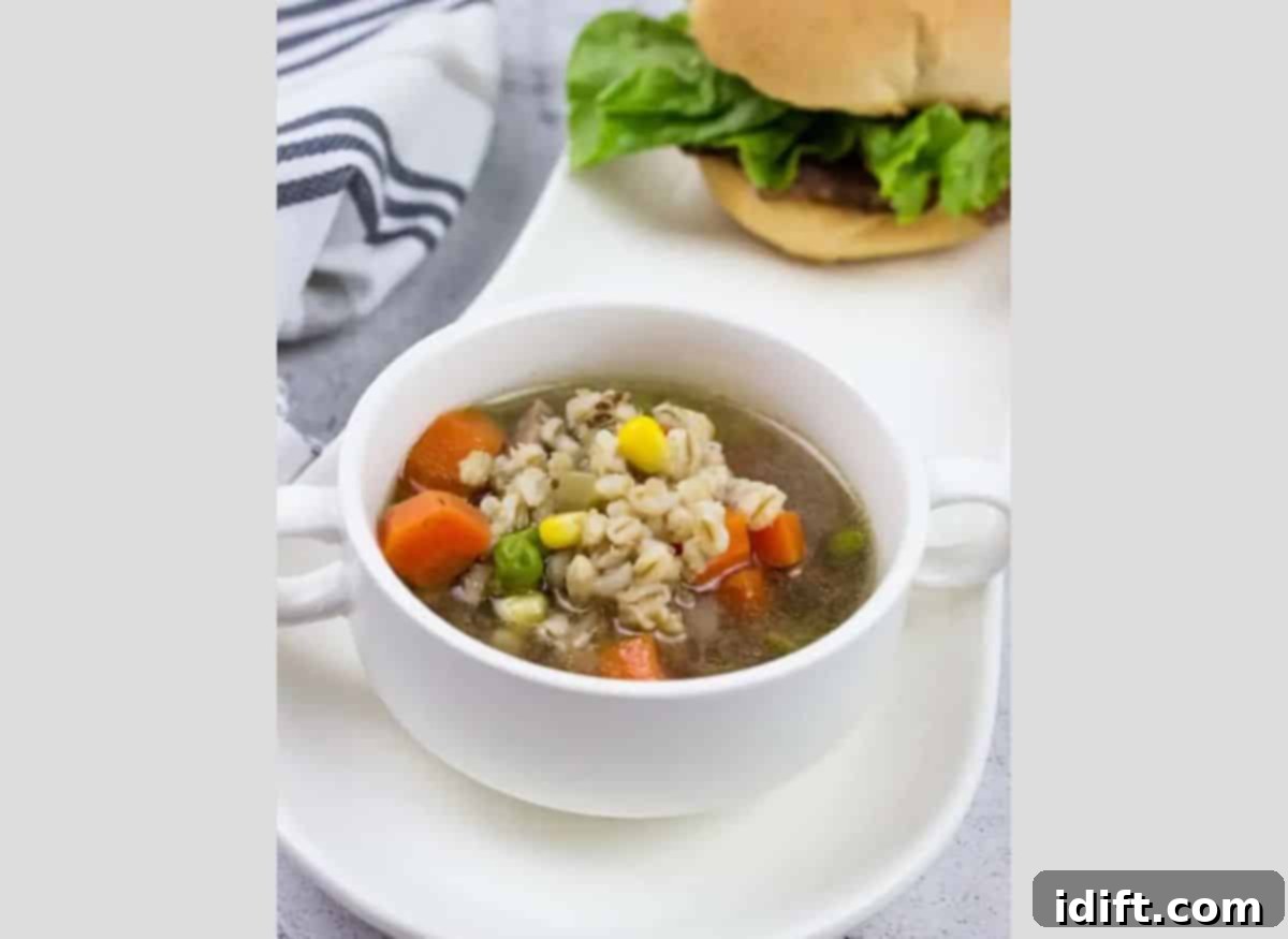 A large bowl of hearty Beef and Barley Soup with a side burger, served on a rustic long white tray.