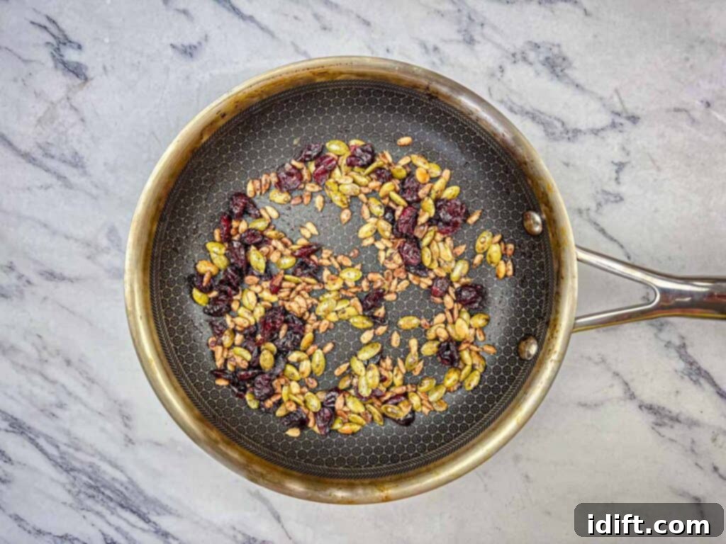 Golden Autumn Salmon Bowl 13 A skillet on medium heat, with pumpkin seeds, sunflower seeds, pumpkin spice, salt, and dried cranberries being stirred together, toasting to a golden crisp for the Harvest Crunch Topping.