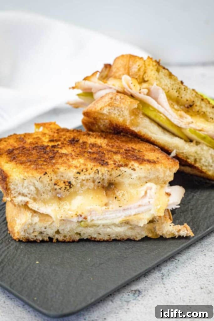 Two halves of a gourmet Turkey & Apple Grilled Cheese sandwich on a slate plate, showcasing melted cheese, smoked turkey, and green apple slices.