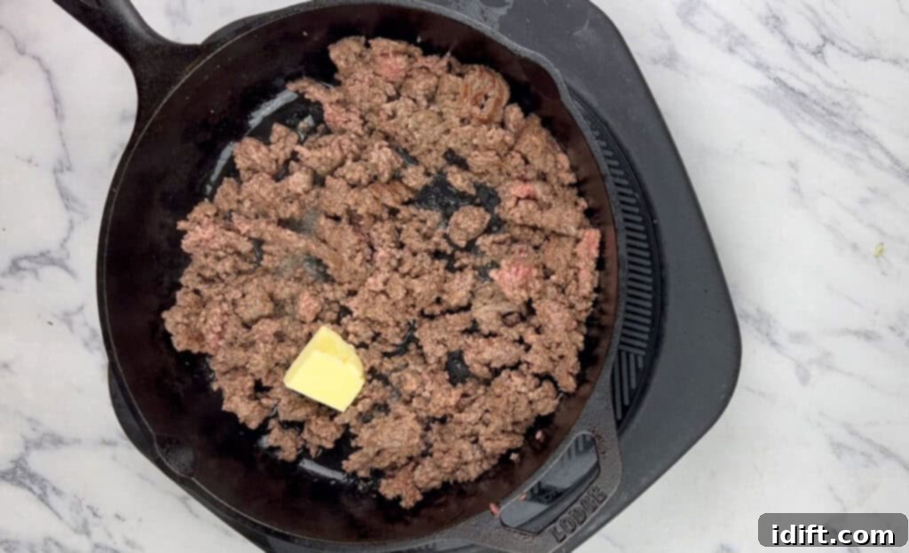 Drained ground beef in a skillet with butter being added.