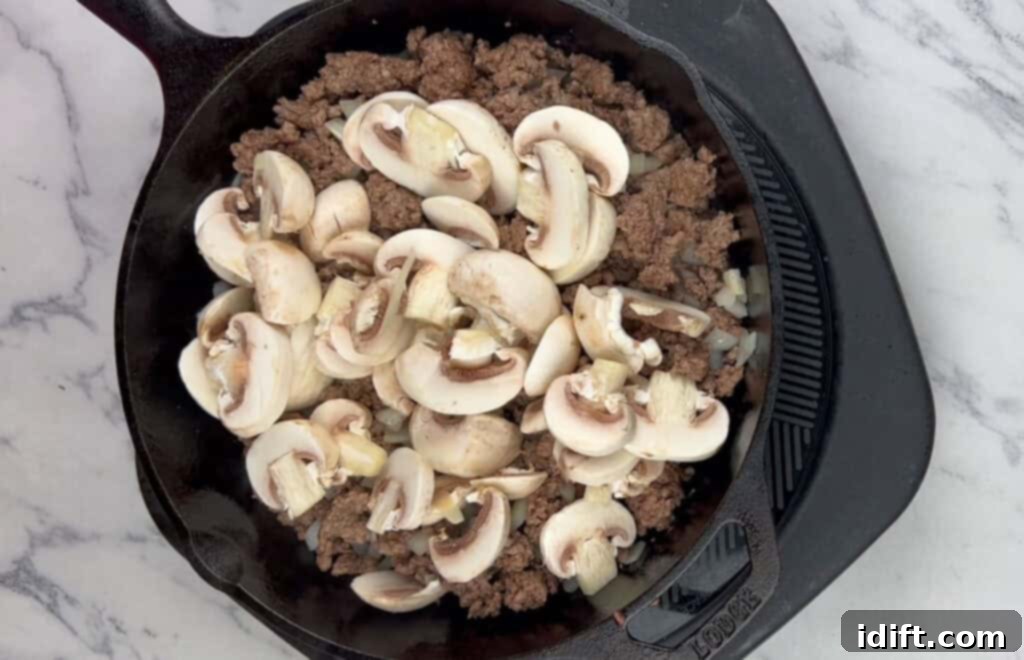Sliced mushrooms being added to the beef and onion mixture in the skillet.