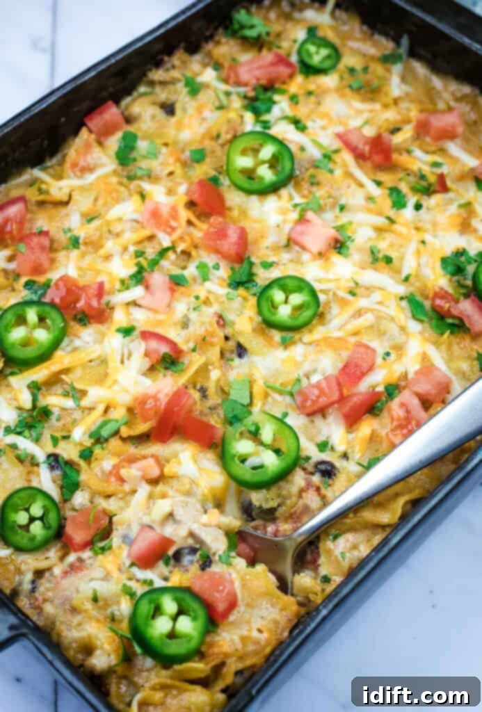 A Cheesy Mexican Casserole topped with melted cheese, jalapeño slices, diced tomatoes, and chopped herbs, with a serving spoon placed on the side.