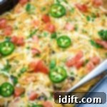 A Cheesy Mexican Casserole topped with melted cheese, jalapeño slices, diced tomatoes, and chopped herbs, with a serving spoon placed on the side.