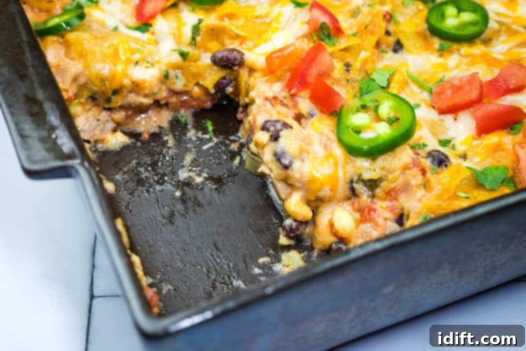 A Cheesy Mexican Casserole in a black dish, featuring layers of cheese, black beans, corn, and topped with jalapeño slices and diced tomatoes. A portion is missing from the corner.