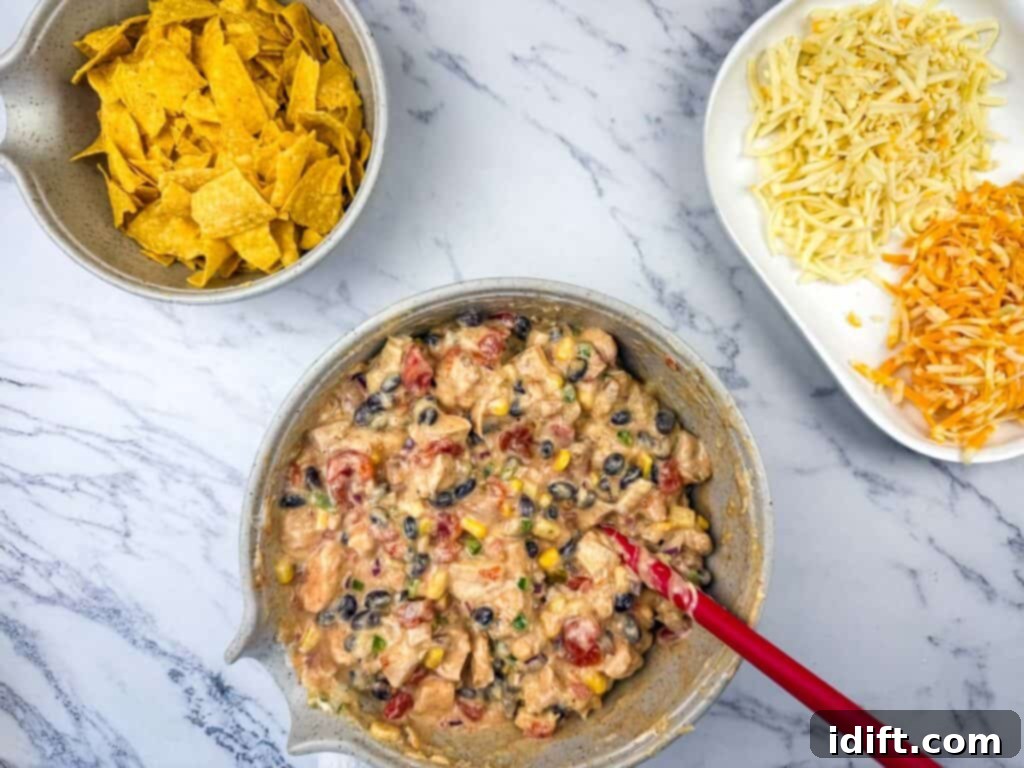 A bowl of creamy dip with tomatoes, black beans, corn, and meat, next to a bowl of tortilla chips and a plate of shredded cheese on a marble surface.