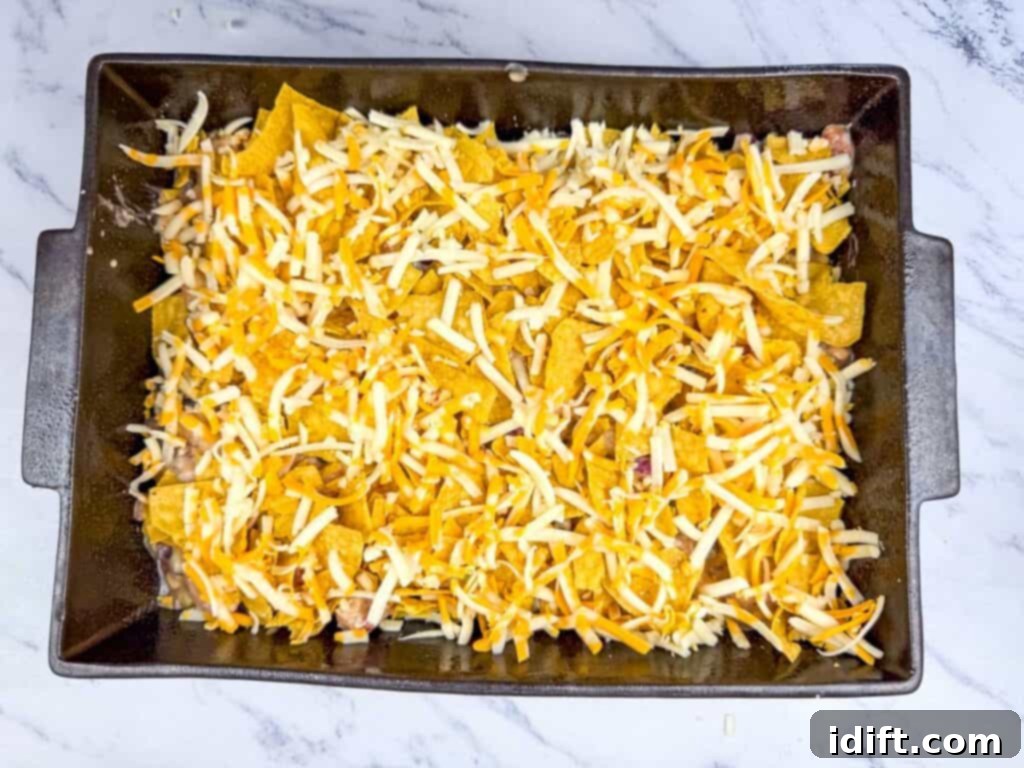 A baking dish filled with tortilla chips, covered with shredded cheddar and mozzarella cheese, on a marble countertop.