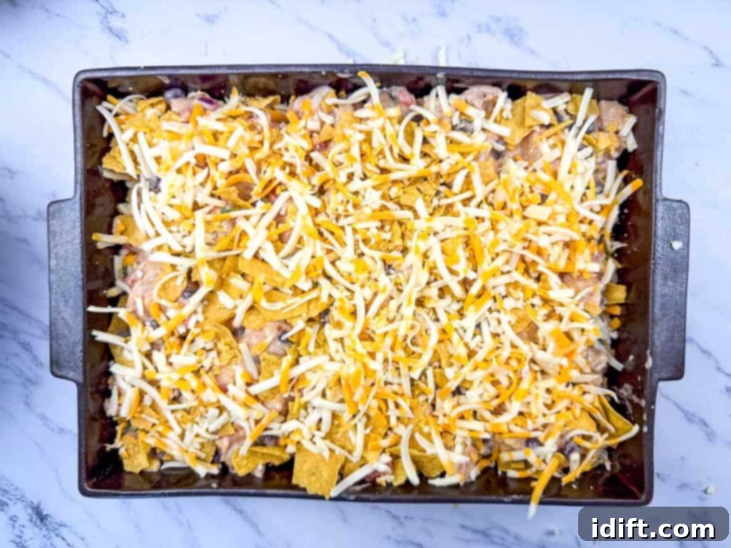 Baking dish with uncooked casserole of shredded cheese, crumbled tortilla chips, beans, and other ingredients on a marble countertop.