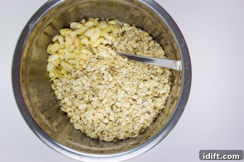 A large metal bowl holds a mixture of rolled oats and sliced almonds, with a metal spoon inside, demonstrating the initial combination of dry ingredients.