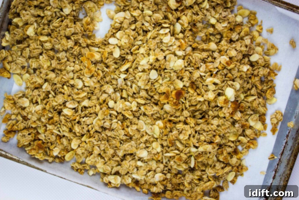 A baking sheet lined with parchment paper, holding a thin, even layer of granola mixture that is partially baked, showing the oats and almonds lightly toasted.