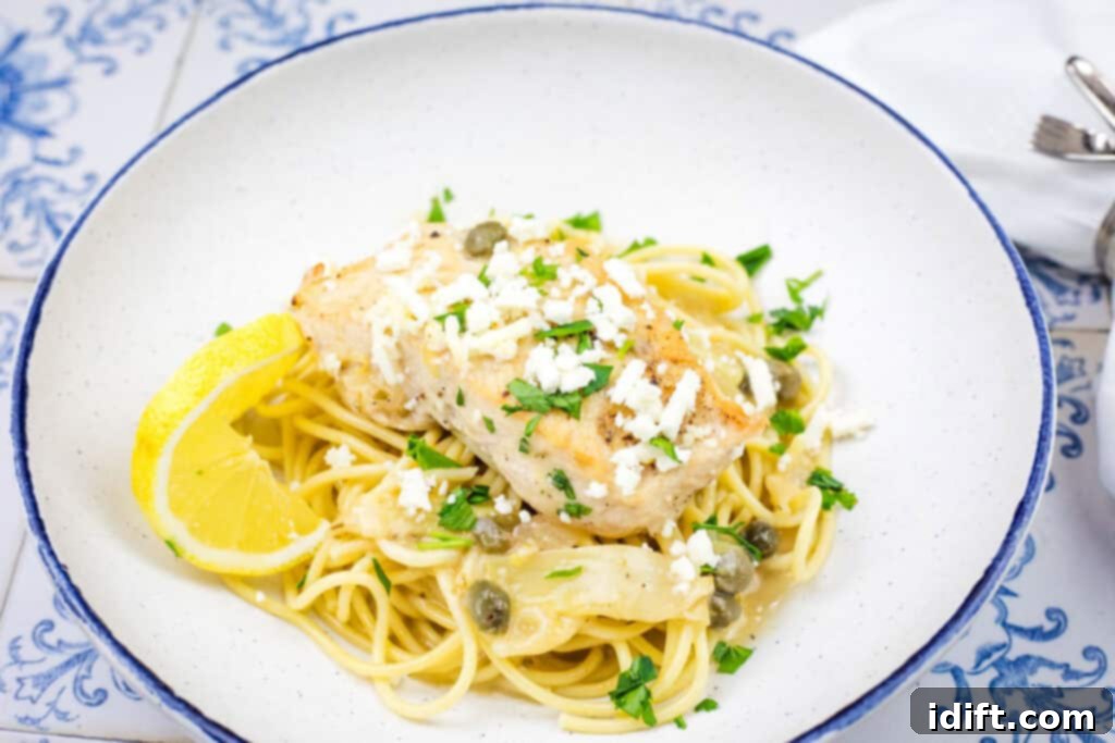 A delectable plate of pasta topped with a piece of Skillet Chicken, generously sprinkled with crumbled feta cheese and fresh herbs, and garnished with a lemon wedge and capers.