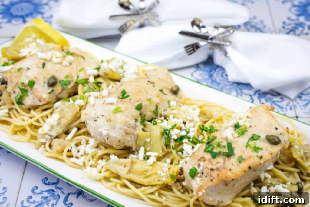 A close-up shot of Skillet Chicken with Artichokes, Lemon, and Feta served with pasta, garnished with fresh capers, chopped parsley, and a sprinkle of cheese. Lemon wedges are arranged on the side, with decorative napkins in the background.