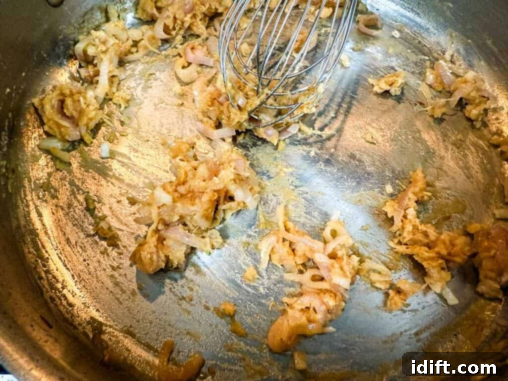 Onions and garlic being stirred with a whisk in a stainless steel pan, as flour is added to form a paste, followed by slowly whisking in chicken broth to create a smooth sauce.