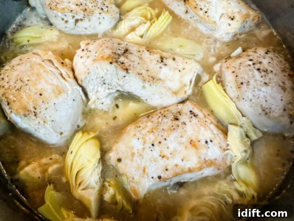 Several seasoned chicken breasts and tender artichoke pieces simmering gently in a flavorful broth, infused with oregano, lemon juice, and zest.