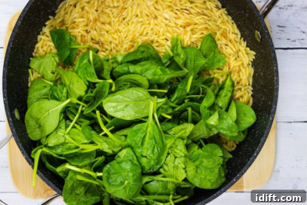 Velvety Spinach and Tomato Orzo 11 Fresh raw spinach being folded into the hot, cooked orzo pasta in a skillet, beginning to wilt.