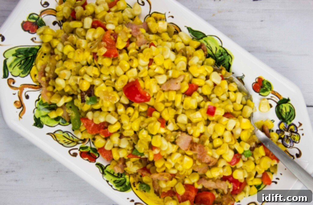 A rectangular dish filled with a corn salad mixed with red bell peppers and small pieces of bacon, with a serving spoon on the side.