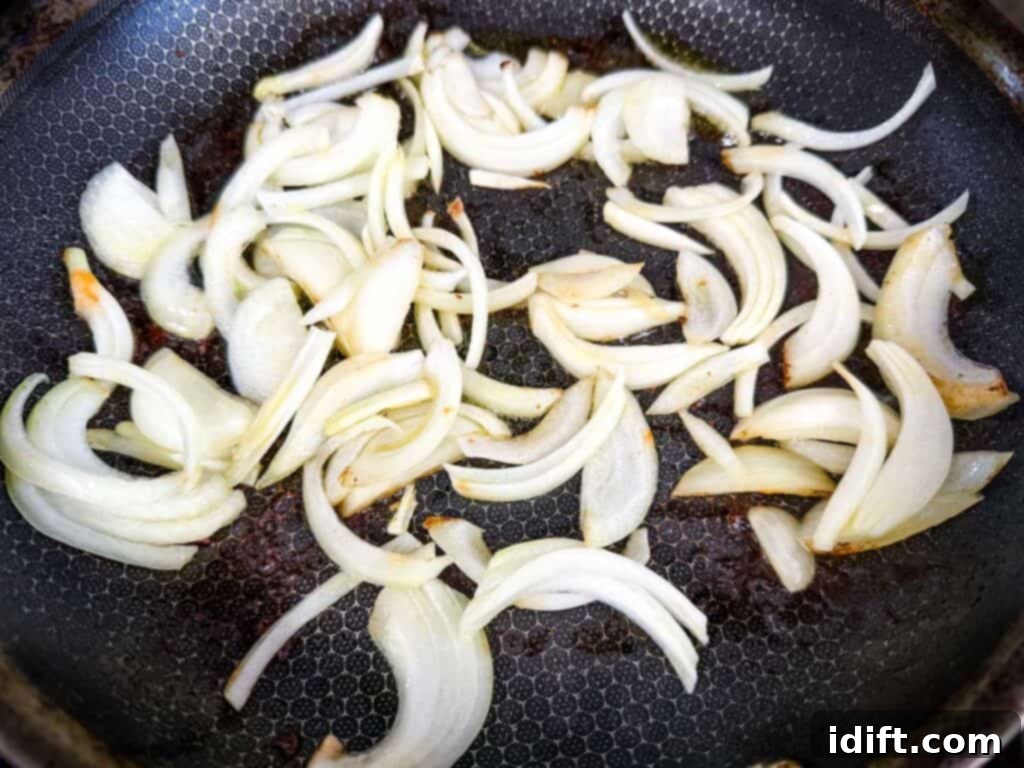 Sliced onions cooking in a frying pan, partially caramelized, releasing their sweet aroma after browning the sausages.