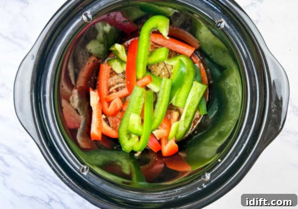 Sliced red and green bell peppers on top of sausages in a slow cooker on a white marble surface, forming layers of flavor.
