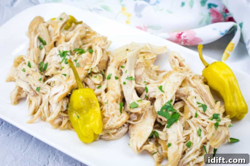 Slow Cooker Mississippi Chicken with chopped herbs and two yellow peppers on a white rectangular plate, placed on a gray surface with a floral cloth in the background.