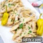 Slow Cooker Mississippi Chicken with herbs and two yellow peppers on a white rectangular plate, placed on a light surface with a floral cloth in the background.