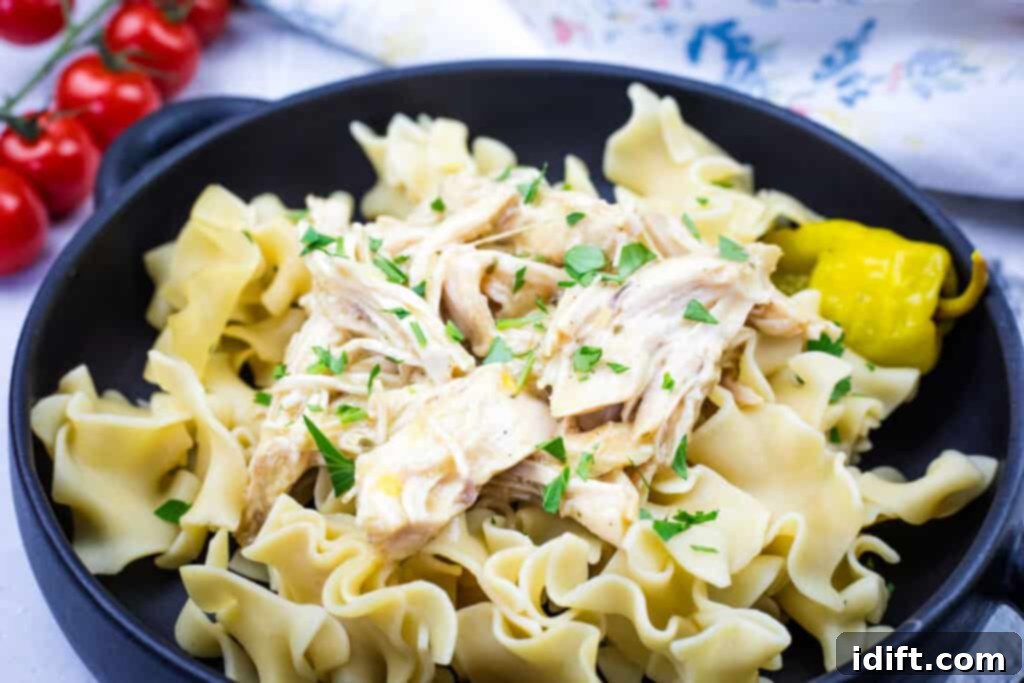 A black bowl containing egg noodles topped with Slow Cooker Mississippi Chicken and garnished with fresh parsley, accompanied by a pickled pepper. Fresh cherry tomatoes are blurred in the background.
