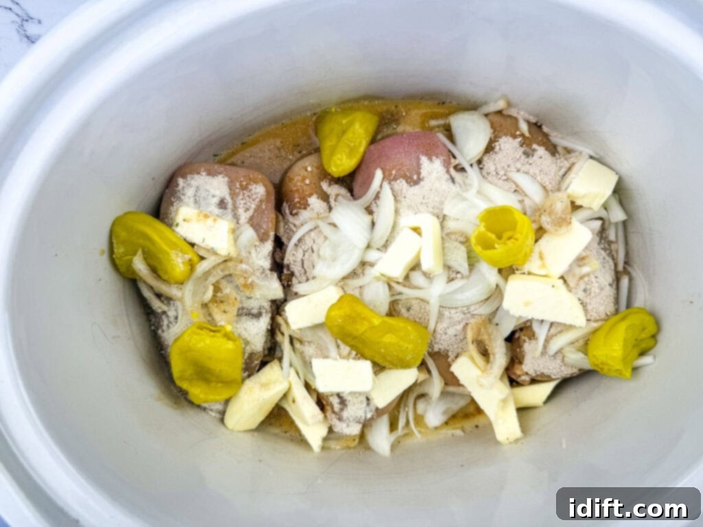 Raw chicken in a slow cooker with sliced onions, butter, pepperoncini peppers, and seasoning.
