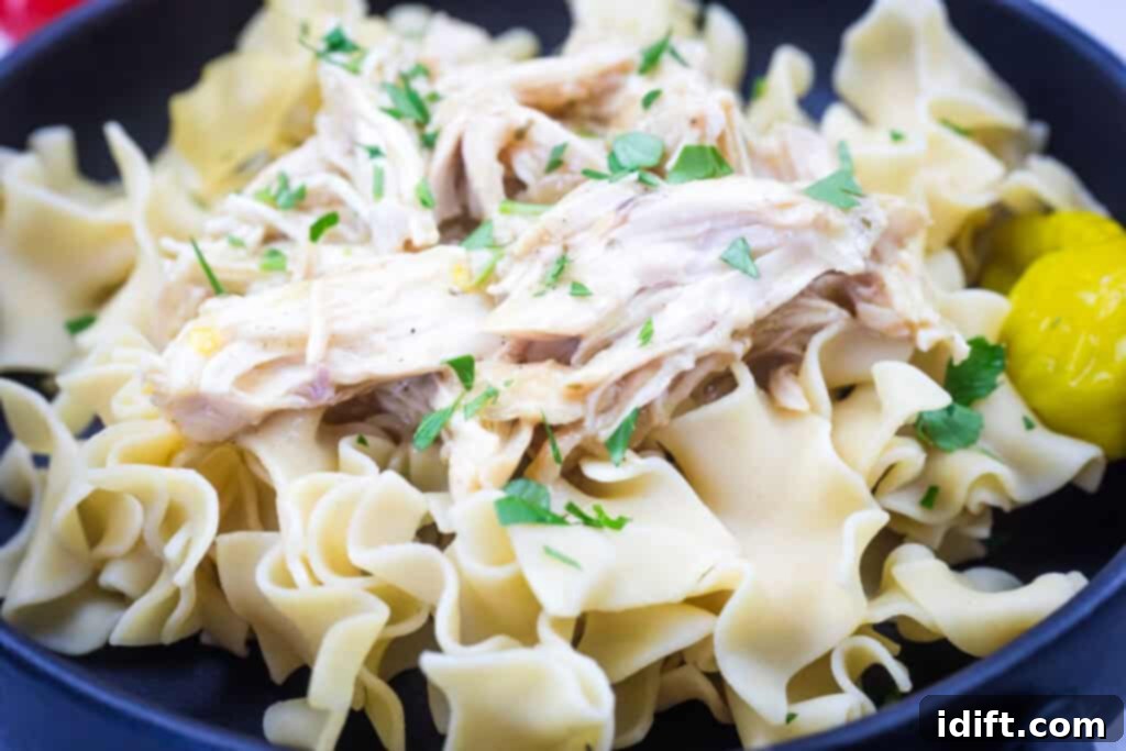 A dish of egg noodles topped with Slow Cooker Mississippi Chicken and parsley, served with a pepper.
