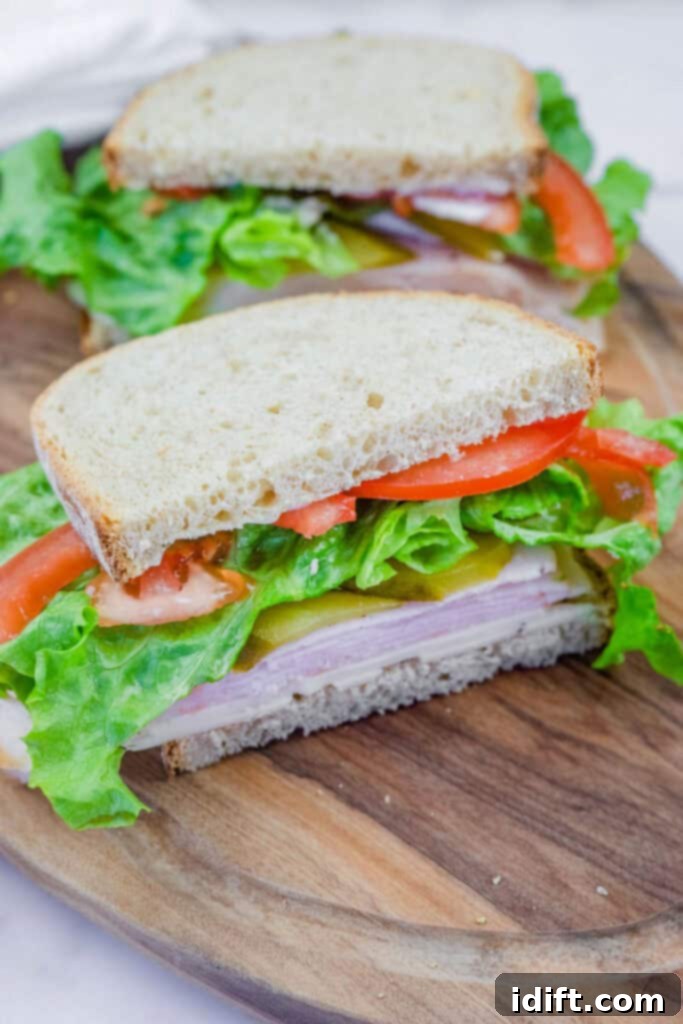 A freshly made turkey sandwich on whole-grain bread, layered with lettuce, tomato slices, pickles, and cheese, served on a wooden cutting board. The sandwich is cut in half, revealing the colorful and fresh ingredients inside.