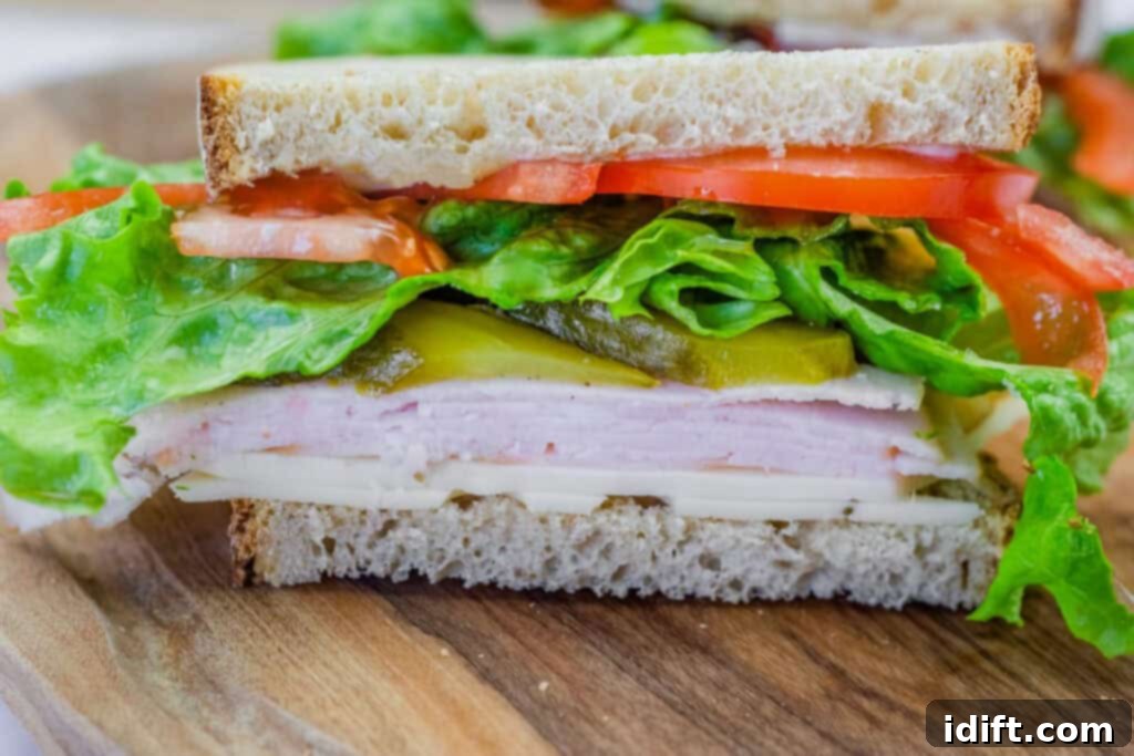 Close-up turkey sandwich with lettuce, tomatoes, pickles, and cheese on whole-grain bread, cut in half and served on a wooden board.