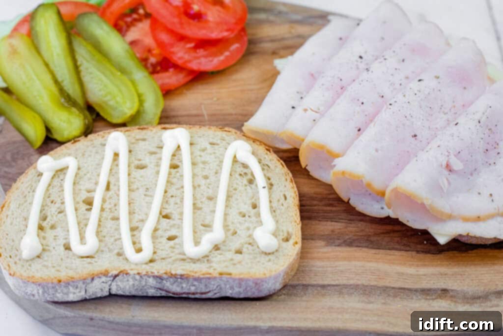 A close-up of sandwich ingredients arranged on a wooden cutting board. A slice of rustic bread with a zigzag drizzle of mayonnaise is in the foreground. To the right, several folded slices of turkey or ham are seasoned with black pepper. In the background, sliced pickles and fresh tomato slices are neatly arranged.