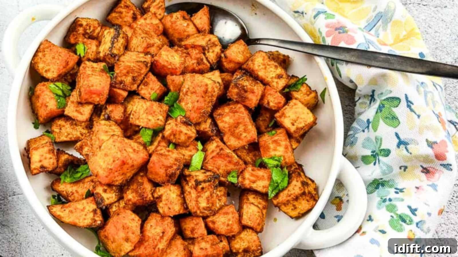 17 Effortless Sides That Prove Dinner Doesn't Have To Be Hard 5 A white bowl filled with seasoned roasted sweet potato cubes, garnished with chopped herbs, with a metal serving spoon on the side and a printed napkin partially visible.