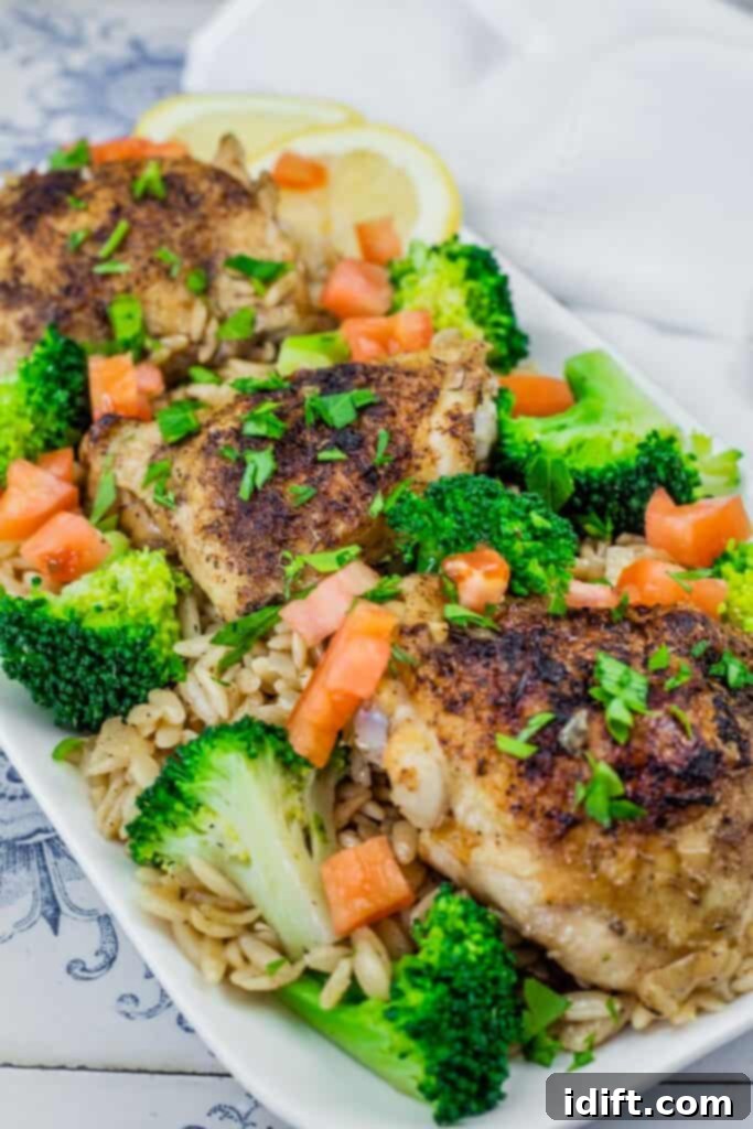 A Lemon Chicken Orzo Skillet with Broccoli dish featuring golden-brown seasoned chicken thighs on a bed of orzo pasta, garnished with fresh broccoli florets, diced tomatoes, chopped parsley, and lemon slices in the background. This one-pan meal is visually appealing and healthy.