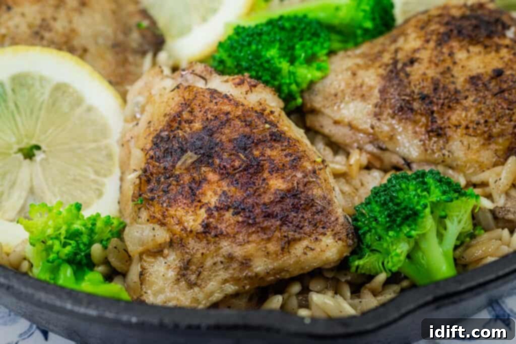 Close-up of crispy, golden-browned lemon chicken thigh atop a bed of orzo pasta, garnished with fresh broccoli florets and lemon slices in a cast-iron skillet, ready for serving.