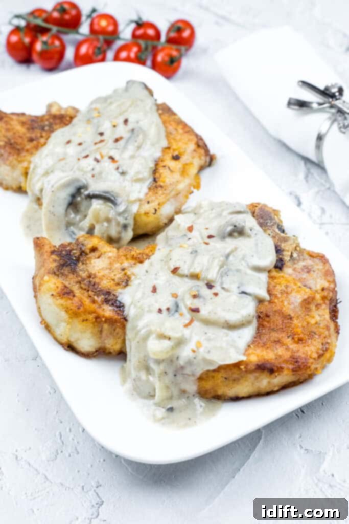 Two Smothered Pork Chops with Onions & Mushrooms on a white plate. Cherry tomatoes and a napkin with a silver holder are in the background.
