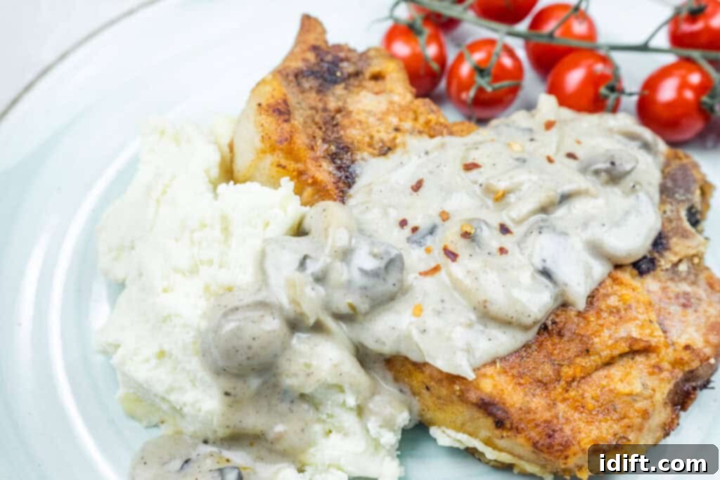 Plate of Smothered Pork Chops with Onions & Mushrooms, served with mashed potatoes and a side of cherry tomatoes.
