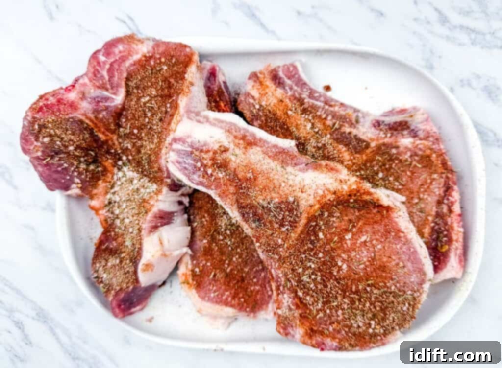 Three raw, seasoned pork chops on a white plate placed on a marble surface.