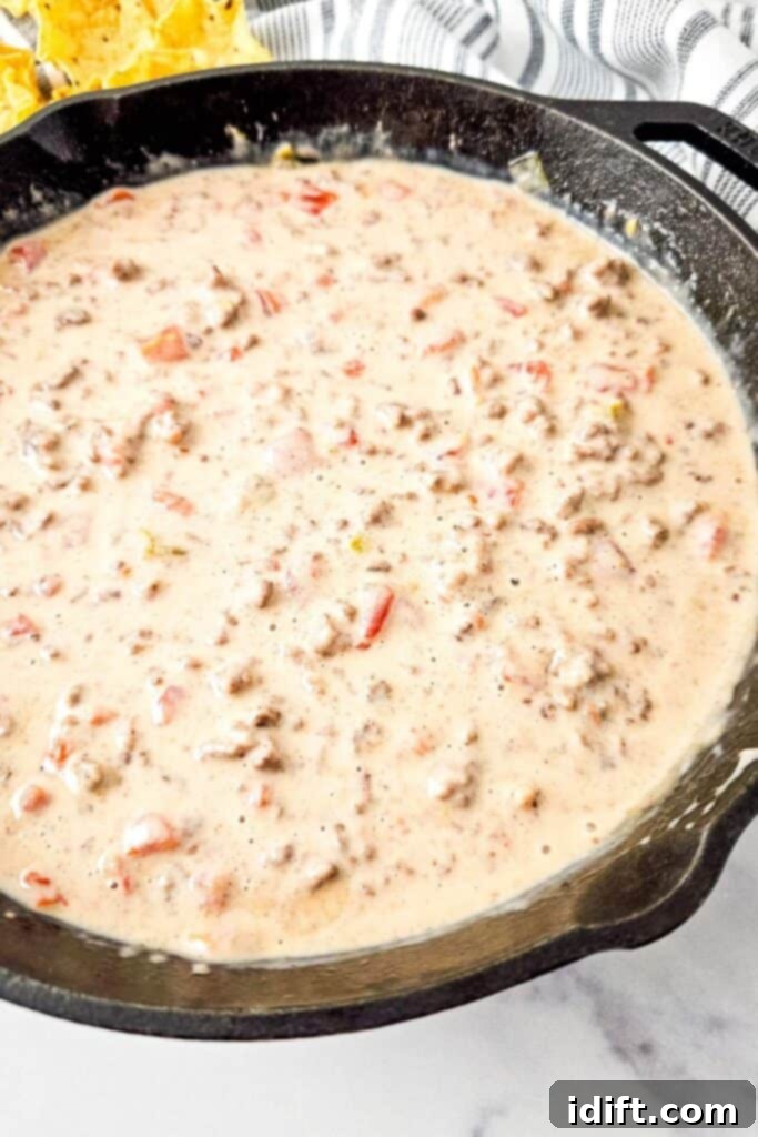 A cast iron skillet filled with creamy Queso Blanco Rotel Dip containing ground meat and diced tomatoes, garnished with fresh cilantro, ready to be served at a party.