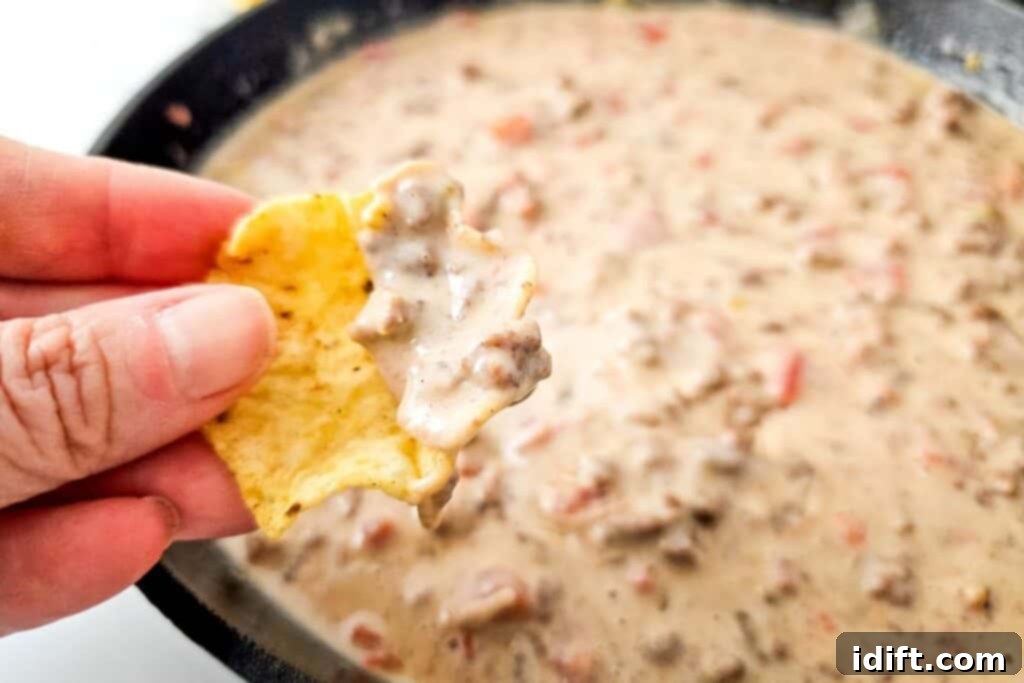 A hand holding a golden tortilla chip, lavishly dipped into the rich, creamy Queso Blanco Rotel Dip directly from a hot cast iron pan, ready to be enjoyed.