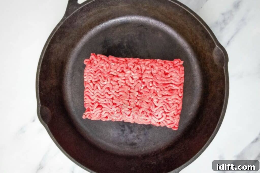 Raw ground beef being added to a hot black cast iron skillet on a marble countertop, ready for the initial browning process.