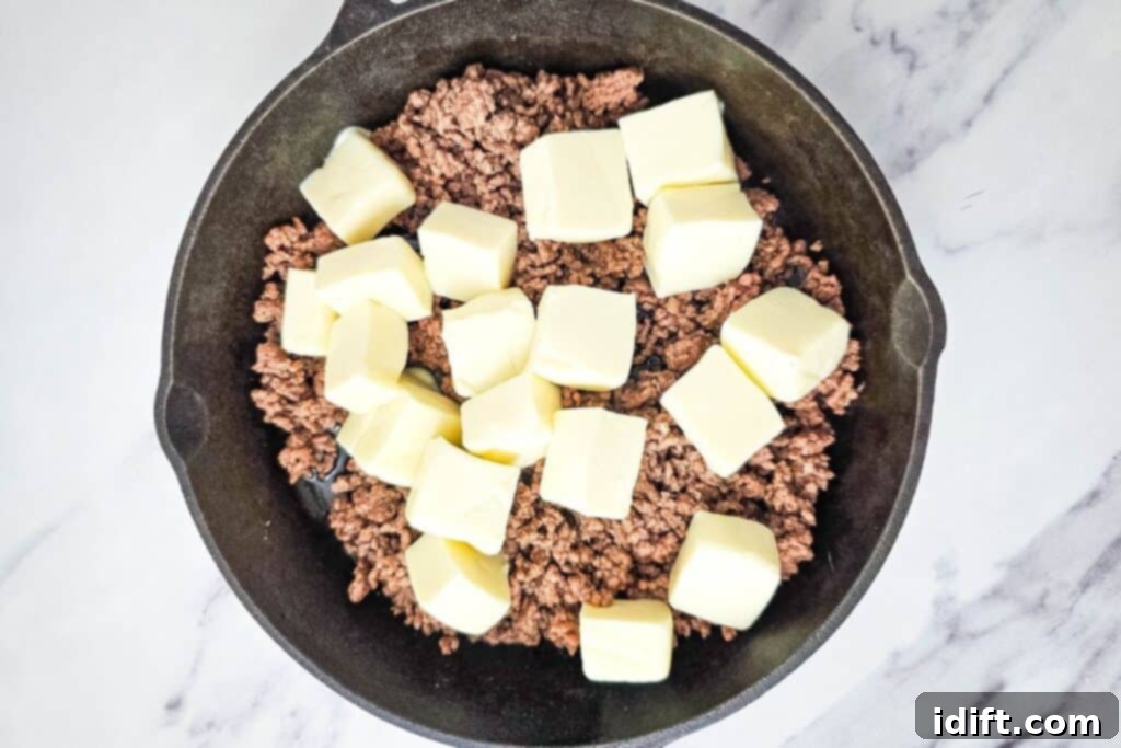 Cooked ground meat mixed with bright orange Velveeta Queso Blanco cheese cubes in a black skillet on a marble surface, beginning to melt and blend.