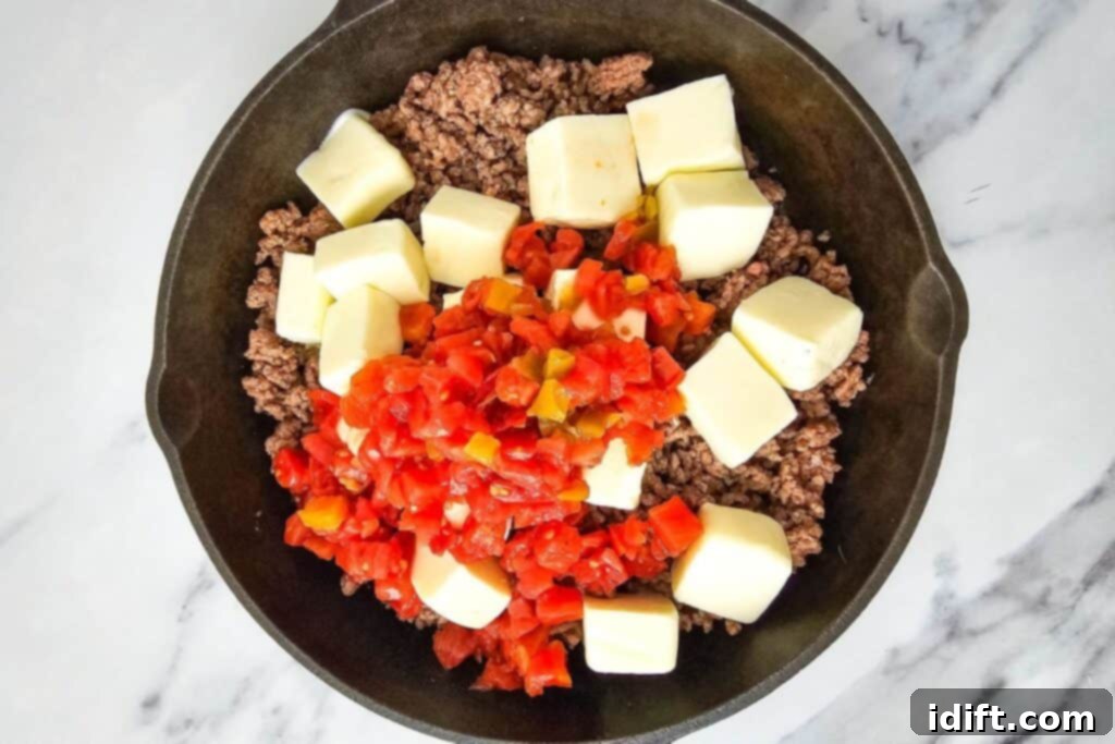 A skillet containing cooked ground beef, diced Rotel tomatoes with green chiles, and several cubes of Velveeta cheese on a marble surface, being stirred to melt into a smooth, creamy dip.