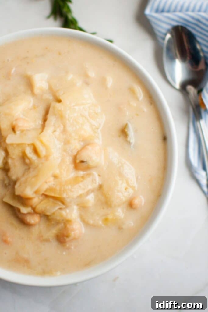 A steaming bowl of Cabbage and Bean Soup, rich with chunks of cabbage and chickpeas, artfully presented on a polished marble surface alongside a rustic wooden spoon and a striped linen napkin, ready to be enjoyed.
