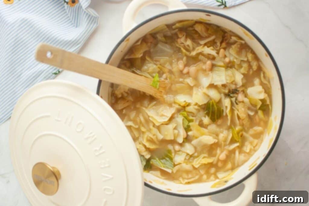 A rustic pot of steaming vegetable soup, featuring tender cabbage, creamy white beans, and vibrant greens, accompanied by a natural wooden spoon. The pot lid rests casually on the side of a polished marble surface, adding to the homely scene.