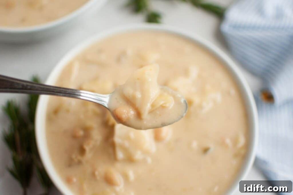 A spoonful of rich, chunky Cabbage and Bean Soup is elegantly held over a brimming bowl, showcasing the tender beans and vegetables. A subtle hint of a striped cloth is visible to the right, adding a touch of domestic warmth.