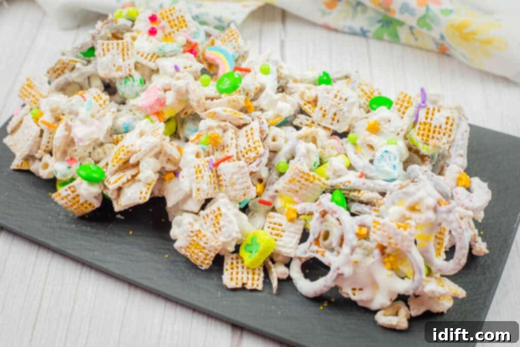 Emerald Enticement 9 A platter of white chocolate-coated snack mix with cereal, pretzels, and colorful candy on a slate board, set atop a white wooden surface.