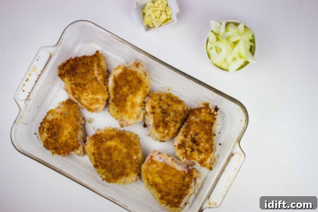 Golden-brown chicken thighs arranged in a single layer in a casserole dish, with sliced onion and garlic scattered around them, ready for baking.