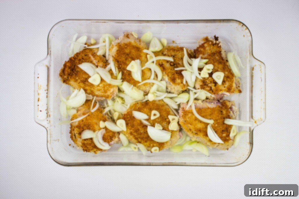 The browned chicken thighs in the baking dish are now topped with a generous scattering of freshly sliced onion and garlic, ready to be covered and baked.