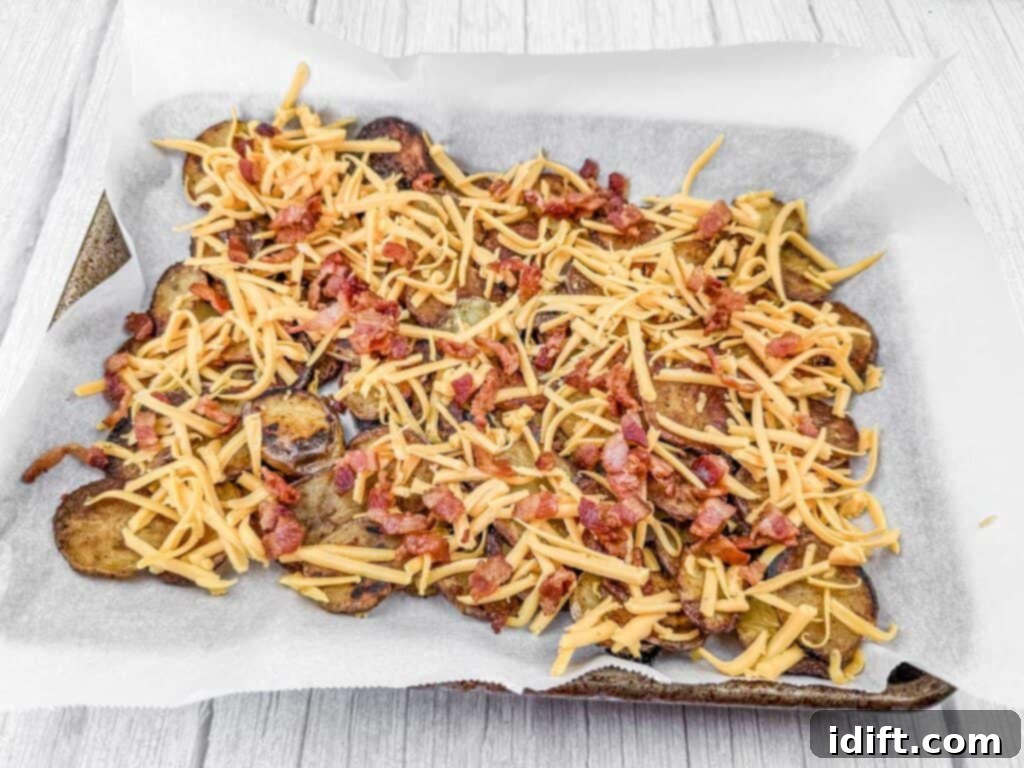 Baked potato slices on a parchment-lined tray, topped with shredded cheese and bacon bits.