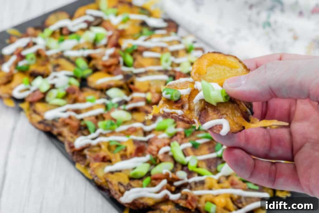 Hand holding a loaded Irish nachos with cheese, bacon, sour cream, and green onions, over a platter of similarly topped slices on a table.
